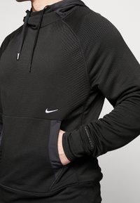 Czarna bluza Nike z materiałem o fakturowanej powierzchni, z kapturem, kangurzą kieszenią i elastycznymi mankietami. Logo na klatce piersiowej i napis "THERMA-FIT" na rękawie.