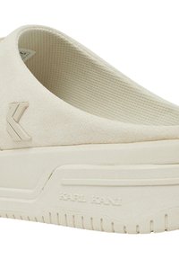 Beige slip-on sko med et teksturert stoffoverdel, en tykk gummisåle og en diskré preget logo detalj på siden.