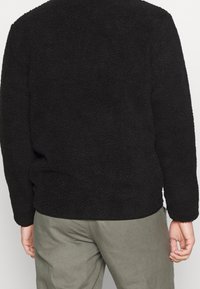 Sudadera de forro polar negra con superficie texturizada y suave, con cuello redondo y mangas largas, combinada con pantalones de tela gris claro.