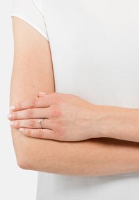 Gull- og sølvring med en liten edelsten, båret på en hånd som hviler på en bar arm, mot en bakgrunn av hvitt stoff. Glatt hudtekstur.