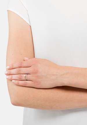 Anello in oro e argento con una piccola pietra preziosa, indossato su una mano appoggiata su un braccio scoperto, su uno sfondo di tessuto bianco. Texture della pelle liscia.
