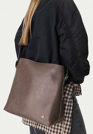 Persona con chaqueta negra y camisa de cuadros lleva un gran bolso de cuero marrón con un pequeño logo metálico en el hombro.