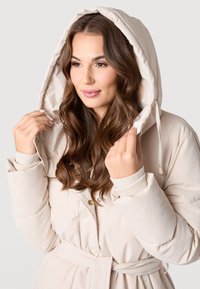 Beige prošívaný kabát s kapucí na stáhnutí, zlatou kovovou sponou, žebrovanými manžetami, páskem v oblasti pasu a hladkým povrchem. Modelka drží kapuci.