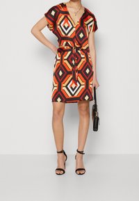 Úžasné šaty s barevným geometrickým potiskem, krátkými rukávy, výstřihem do V, s páskem v pase a přiléhavým střihem. Doplňuje je černé sandály na podpatku a kabelka.