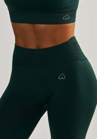 Ensemble de vêtements de sport vert foncé comprenant un crop top ajusté et un legging taille haute, tous deux avec une texture lisse et un logo en forme de cœur en blanc.