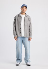 Light gray button-up shirt with a pocket, white t-shirt underneath, light blue baggy jeans, and a blue baseball cap. Sneakers complete the look.