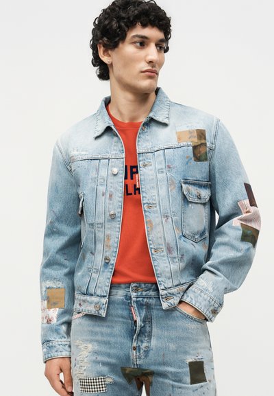 Νέος άνδρας με ανοιχτό μπλε τζιν μπουφάν και τζιν με λεπτομέρειες patchwork, σε συνδυασμό με κόκκινο γραφικό T-shirt, στέκεται μπροστά σε απλό φόντο.