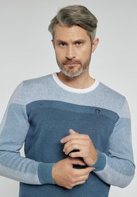 Niebieski sweter z dzianiny z gradientowym wzorem w różnych odcieniach niebieskiego, białym dekoltem i małym logo na klatce piersiowej. Długie rękawy z ściągaczami.