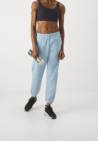 Námořnicky modrá sportovní podprsenka, světle modré tepláky s elastickými manžetami a černé tenisky. Drží stříbrnou činku s hladkou texturou.