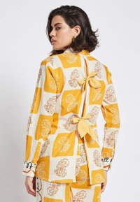 Yellow and cream patterned jacket featuring loose bows at the back, long sleeves, and a straight cut. Fabric texture appears smooth.