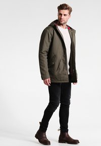 Parka vert olive avec une capuche, deux poches avant et doublure en tartan rouge. Modèle portant un jean noir et des bottines brunes.