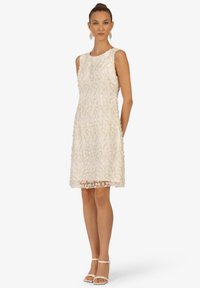 Ärmeloses, floral besticktes Kleid in Cremefarbe. Es hat eine figurbetonte Silhouette und Spitzenverzierungen am Saum, kombiniert mit Riemchensandalen.
