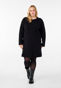 Svart stickad klänning med långa ärmar och rundad halsringning, med rak skärning och sidofickor, tillsammans med svarta knähöga stövlar.