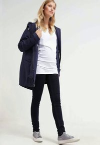 Parka azul-marinho com capuz, zíperes frontais e bolsos, sobre uma t-shirt branca, combinada com jeans skinny escuros e ténis cinzas.