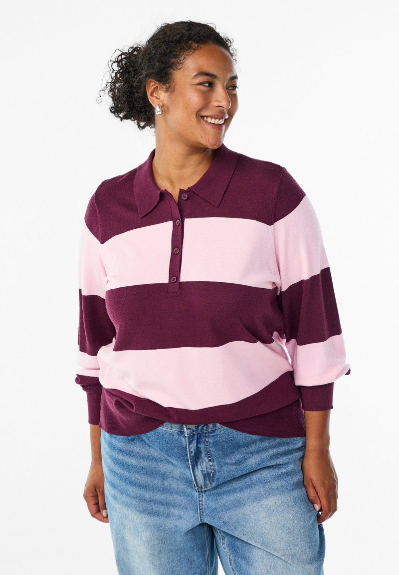 Bordeaux- og lyserødstribet polo-sweater med krave og knaplukning. Har lange ærmer med opbrettede manchetter. Båret med lyseblå jeans.