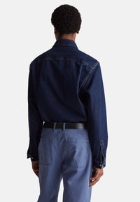 Camicia di jeans blu scuro con maniche lunghe e colletto a punta, abbinata a pantaloni blu chiaro. Cintura nera visibile. Design semplice e strutturato.