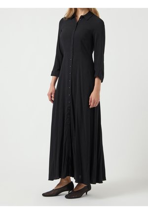 Frau trägt ein langes schwarzes Hemdkleid mit dreiviertel Ärmeln und schwarzen niedrighackigen Schuhen, steht vor einem einfarbigen Hintergrund.