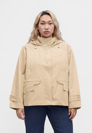Person med langt blond hår iført en beige hættetrøje med forlommer og blå jeans mod en ensfarvet baggrund.