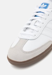 Vit sneaker med accenter i grått ruggat läder, med rund tå, strukturerad brun gummisula och en blå logotag på tungan.