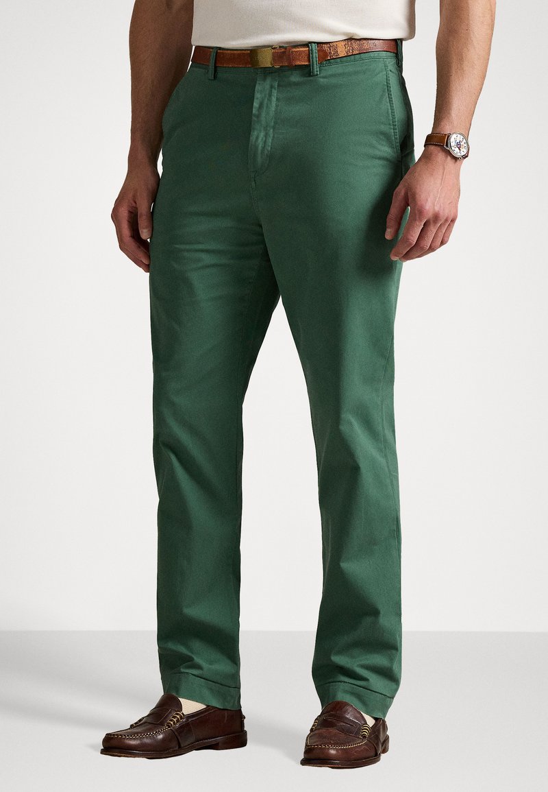 Zaļie kokvilnas chinos bikses ar taisnu piegriezumu, sānu kabatām un gludu tekstūru. Valkātas ar brūnu ādas jostu un brūniem mokasīniem.