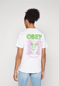 Balta medvilninė marškinėliai su dideliu žaliu "OBEY" grafiniu dizainu ir rožinės grandinės garde su veido raštu bei tekstu "BREAKING CHAINS WORLDWIDE".