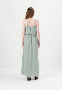 Femme aux cheveux ondulés portant une robe vert clair, sans manches, longueur cheville, avec de petits pois blancs, debout devant un fond blanc uni.