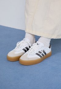 Zapatillas blancas con rayas negras y acentos de ante beige. Cuentan con una suela de goma texturizada y cordones blancos planos, usadas con calcetines de color claro.