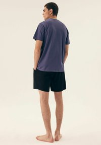 Fioletowa bawełniana koszulka z krótkim rękawem, zestawiona z czarnymi szortami. Model stoi boso, z rękami w kieszeniach, widziany z tyłu.