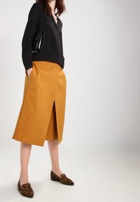 Schwarze V-Ausschnitt-Bluse, senfgelbe Wickelrock und Schuhe mit Leopardenmuster. Der Rock hat einen Schlitz vorne und Taschen und besteht aus glattem Stoff.
