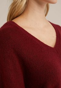 Frau trägt einen burgunderfarbenen, strukturierten V-Ausschnitt-Pullover, der ihren Hals, ihre Schulter und ihr blondes Haar vor einem schlichten Hintergrund zeigt.