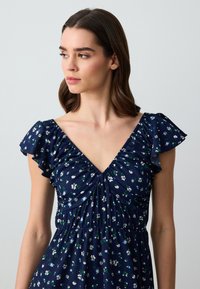 Marineblaues Kleid mit kurzen Rüschensäulen, das einen tiefen V-Ausschnitt und ein florales Muster in Weiß und Grün aufweist. Strukturstoff mit gerafftem Design.