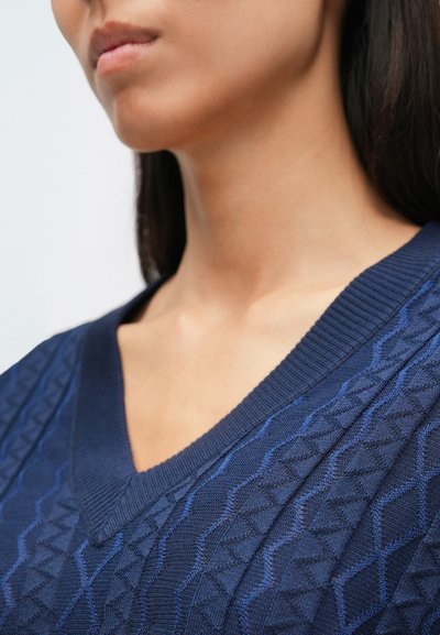 Tamsiai mėlynas mezginys megztinis su faktūrine geometrine raštuote, V formos iškirpte ir dantytais apykaklės kraštais. Minkšta medžiaga su tūrine detalizacija.