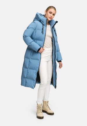 Abrigo largo acolchado en azul claro, con capucha, cremallera frontal y bolsillos laterales. Llevado encima de una camiseta beige y pantalones blancos con botas beige.