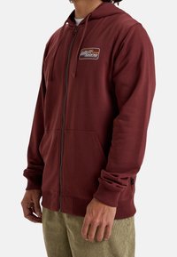 Sudadera con cremallera de color marrón burdeos, con un parche de logo, bolsillos frontales, puños acanalados y una textura suave. Presenta una capucha con cuerda y un corte relajado.