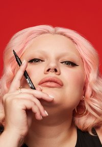 Un bolígrafo de eyeliner negro sostenido por una persona con cabello ondulado rosa y un anillo en el septum, en un fondo rojo brillante. Color de labios neutro y pendientes de aro dorados.