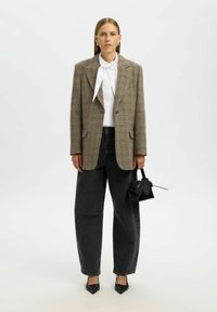 Naine seisab seljas pruunist ruudulisest bleiserist, valgest pluusist koos paelaga, lohvakatest mustadest teksadest ja mustadest teravate ninaga kingadest, hoides väikest musta käekotti valge tausta ees.