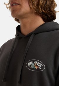 Felpa grigia con cappuccio caratterizzata da un logo "BILLABONG" ricamato con dettagli floreali. Il materiale ha una consistenza morbida e presenta i cordini sul cappuccio.