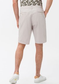 Muž v béžových šortkách a bílých teniskách, pohled zezadu na bílém pozadí.