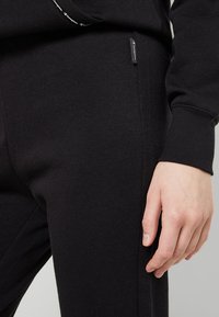 Los pantalones de chándal negros de algodón presentan una textura suave, bolsillos laterales y una pequeña etiqueta con el logo de la marca en la cintura. Mano descansando sobre la tela.