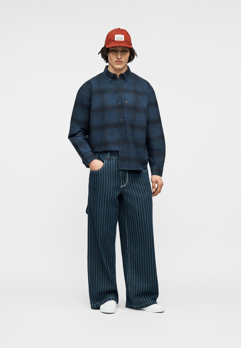 Jeune homme portant une casquette rouge, une chemise à carreaux bleu foncé avec boutons, un pantalon large à fines rayures et des baskets blanches, debout avec une main dans la poche.