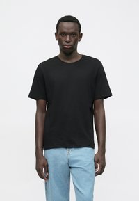 Giovane uomo con capelli corti, che indossa una semplice t-shirt nera e jeans azzurri, in piedi contro uno sfondo bianco semplice.