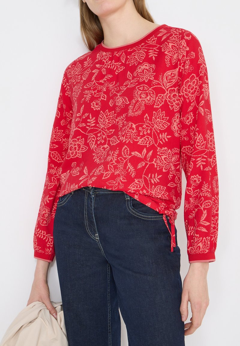 Femme portant un haut à manches longues rouge avec des motifs floraux beiges et un jean bleu foncé, tenant un vêtement beige clair dans sa main gauche.