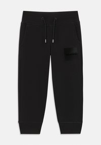 Pantalons de survêtement noirs en tissu doux, dotés d'une taille élastique, d'un cordon de serrage et d'une étiquette logo noire sur la cuisse gauche.