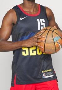 Czarny strój koszykarski z czerwonymi akcentami, z żółtymi i białymi numerami oraz napisami. Zawodnik trzyma pomarańczową piłkę do koszykówki z niebieskimi akcentami.