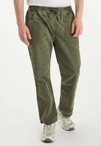 Man in olijfgroene cargobroek met trekkoord in de taille en witte sneakers, staande tegen een effen lichte achtergrond.