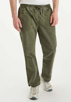 Man in olijfgroene cargobroek met trekkoord in de taille en witte sneakers, staande tegen een effen lichte achtergrond.