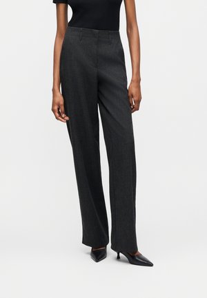 Osoba nosiaca tmavosivé široké nohavice, čierny top a čierne lodičky s ostrou špičkou stojaca na jednoliatom bielom pozadí.