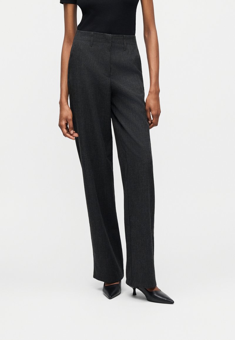 Άτομο που φοράει σκούρα γκρι παντελόνια με φαρδιά πόδια, μαύρο τοπ και μαύρα ψηλοτάκουνα παπούτσια με μυτερή μύτη, στέκεται σε απλό λευκό φόντο.