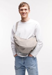 Borsa a tracolla grigia in tessuto con forma curvata, tracolla regolabile e logo sobrio. Il modello indossa una camicia bianca e jeans blu.