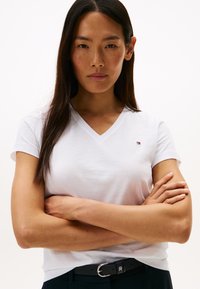 Odrasla ženska oseba z dolgimi temnimi lasmi, oblečena v belo T-majico z V-izrezom in črne hlače s črnim pasom, stoji z rokami prekrižanimi.